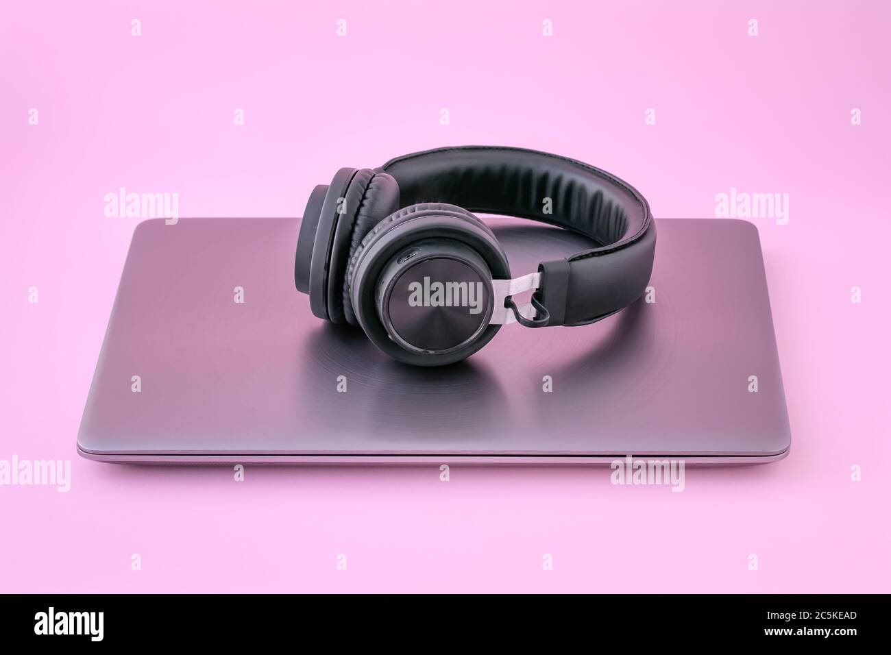 Laptop aus Metall und schwarze Kopfhörer auf rosa Hintergrund. Geschlossenes Notebook mit Ohrhörern. Das Konzept der Selbstbildung Stockfoto