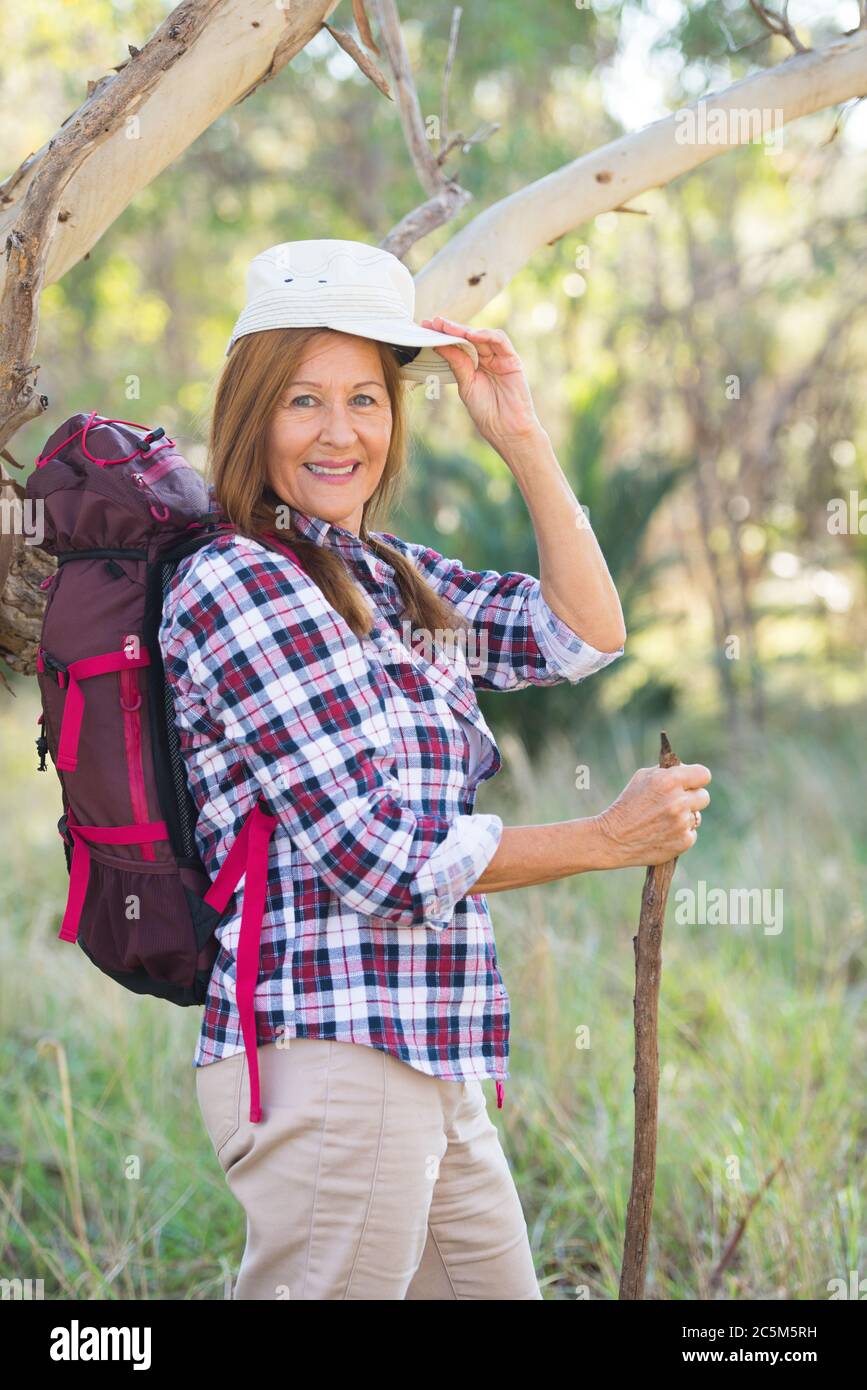 Portrait aktiv Attraktive Senior Frau, die glücklich mit Rucksack und Stock in den Wald, trägt kariertes Hemd und Mütze, verschwommen australischen Hintergrund Stockfoto