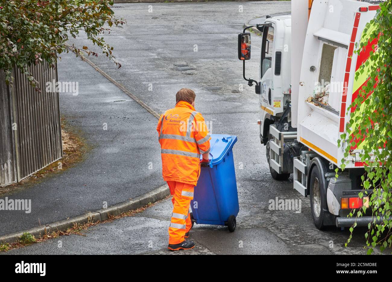 Ein Recycling-Sammler in orangefarbener Kleidung schiebt einen blauen Mülltonnen voller Haushaltsmüll in Richtung eines Müllsammelwagens. Stockfoto