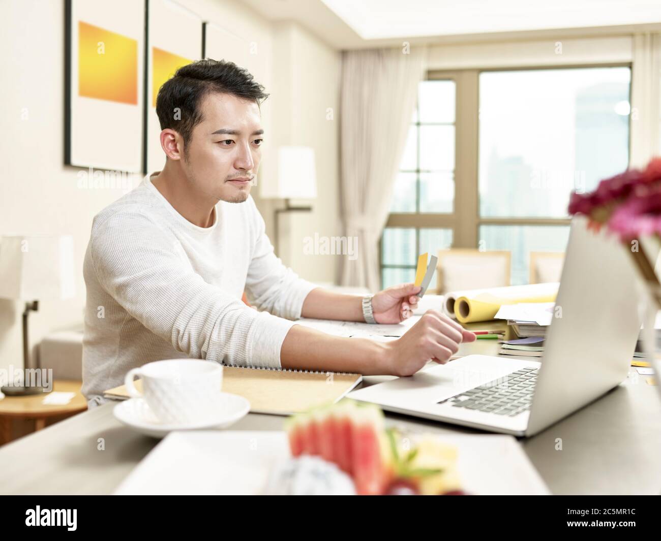 Junge asiatische Mann Design professionelle Arbeit von zu Hause sitzen in der Küche mit Laptop-Computer (Kunstwerk im Hintergrund digital verändert) Stockfoto