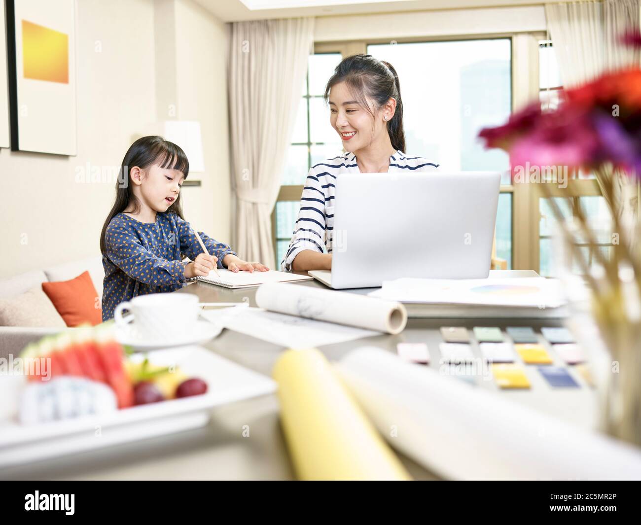 Junge asiatische Designer Mutter arbeitet von zu Hause mit Laptop-Computer während der Betreuung der Tochter (Kunstwerk im Hintergrund digital verändert) Stockfoto
