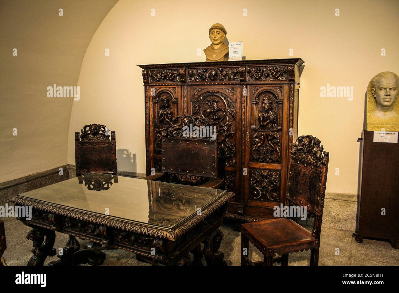 Toledo, Castilla-La Mancha, Spanien, Europa. Kloster San Juan de los Reyes ((14771504). Ein Tisch mit Stühlen, ein Holzmöbel mit Intarsien und zwei Büsten im Vorzimmer des Eingangs zum Kreuzgang. Stockfoto