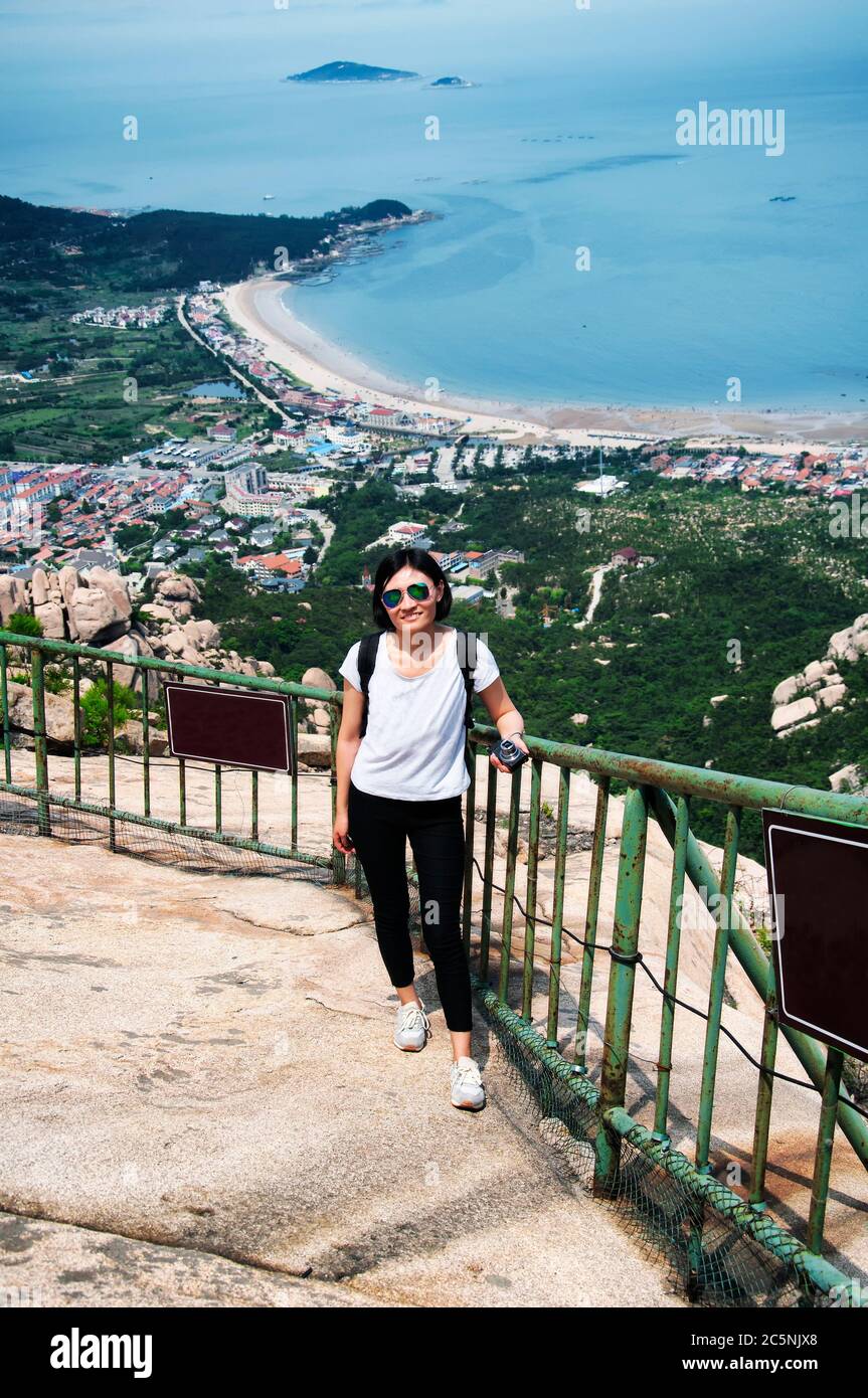 Eine lächelnde chinesin mit Sonnenbrille auf einem Aussichtspunkt auf dem Berg Lao mit dem ostchinesischen Meer im Hintergrund in Qingdao China shandong provin Stockfoto