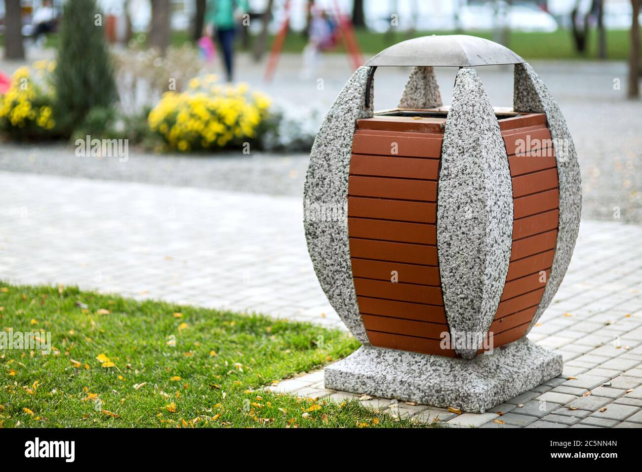 Eine ovale Mülltonne auf einem Betonständer, der mit Brettern vor dem Hintergrund eines Fußgängerwegs eines Parkbereichs dekoriert ist. Stockfoto