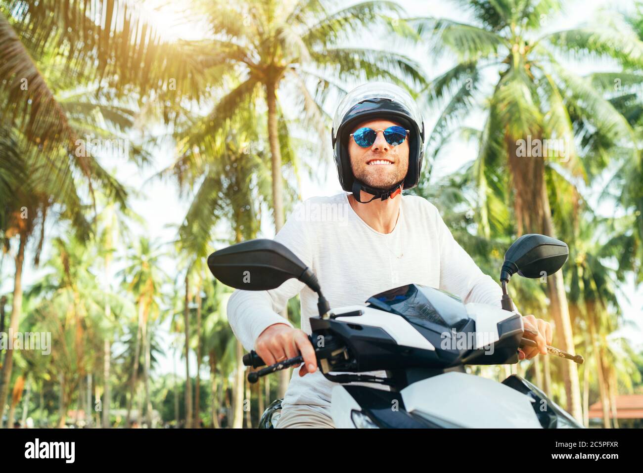 Glücklich lächelnd männlichen Touristen in Helm und Sonnenbrille Reiten Motorrad Roller während seines tropischen Urlaubs unter Palmen. Stockfoto