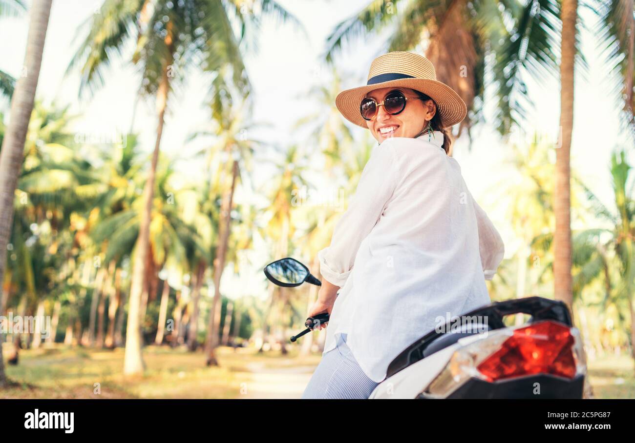 Glücklich lächelnde Frau in Strohhut und Sonnenbrille Reiten Motorrad unter Palmen. Sorgloses Urlaubskonzept. Stockfoto
