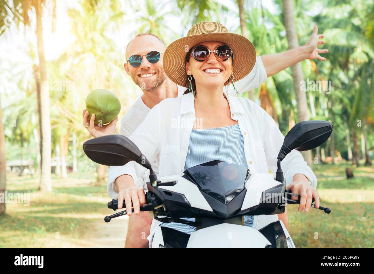 Glücklich lächelnd Paar Reisende Reiten Motorrad Roller unter Palmen. Lachender Mann, der eine Kokosnuss in der Hand trägt.Tropisches Urlaubskonzept Stockfoto