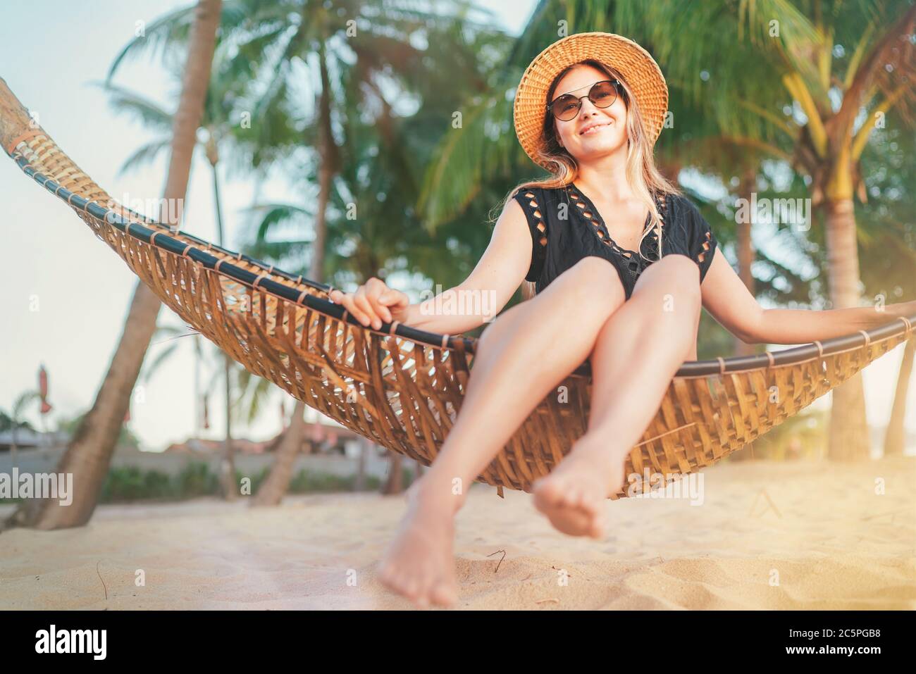 Fröhliche lächelnde junge Frau in Strohhut in Sonnenbrille sitzt in der Hängematte und genießen Sie Morgensonne Licht auf dem Koh Samui tropischen Strand, Thailand. Stockfoto