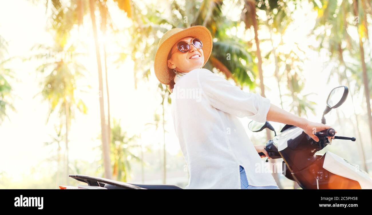 Glücklich lächelnde Frau in Strohhut und Sonnenbrille Reiten Motorrad unter Palmen die Bäume. Sorglos warm Counries Urlaub Zeit Konzept Bild. Stockfoto