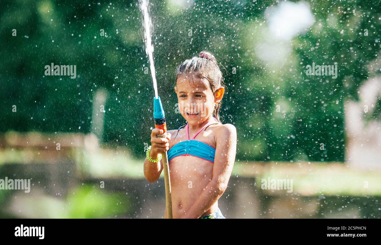 Nettes kleines Mädchen mit Wasser für sich selbst aus dem Schlauch bestreuen, so dass ein regen Vergnügen für heiße Sommertage. Sorgloses Konzept der Kindheit. Stockfoto