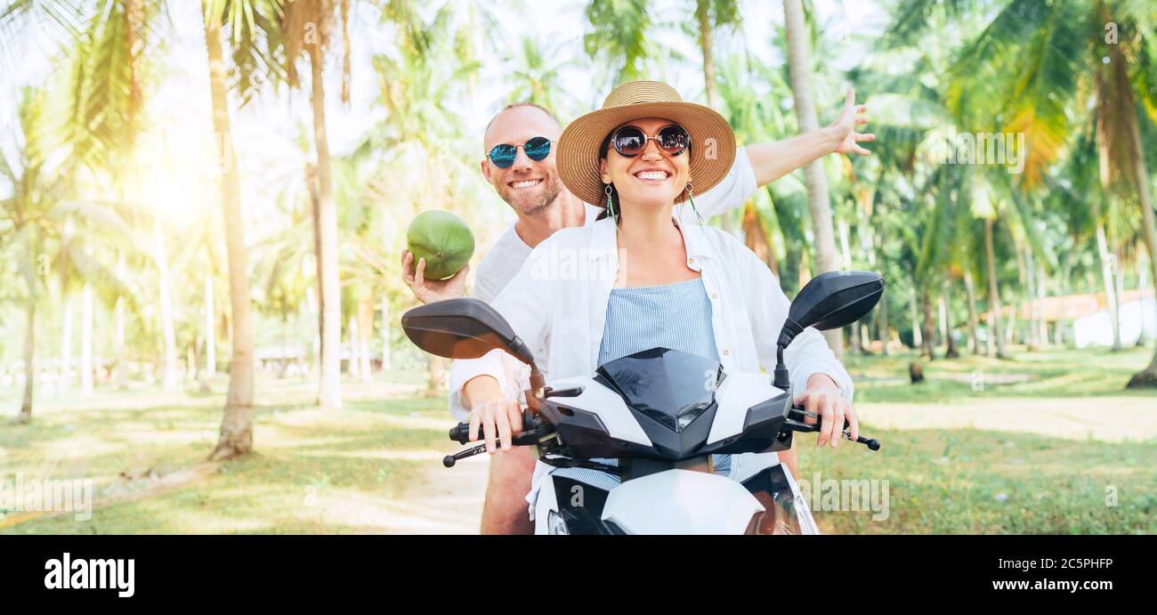 Glücklich lächelnd Paar Reisende Reiten Motorrad Roller unter Palmen. Lachender Mann, der eine Kokosnuss in der Hand trägt.Tropisches Urlaubskonzept Stockfoto