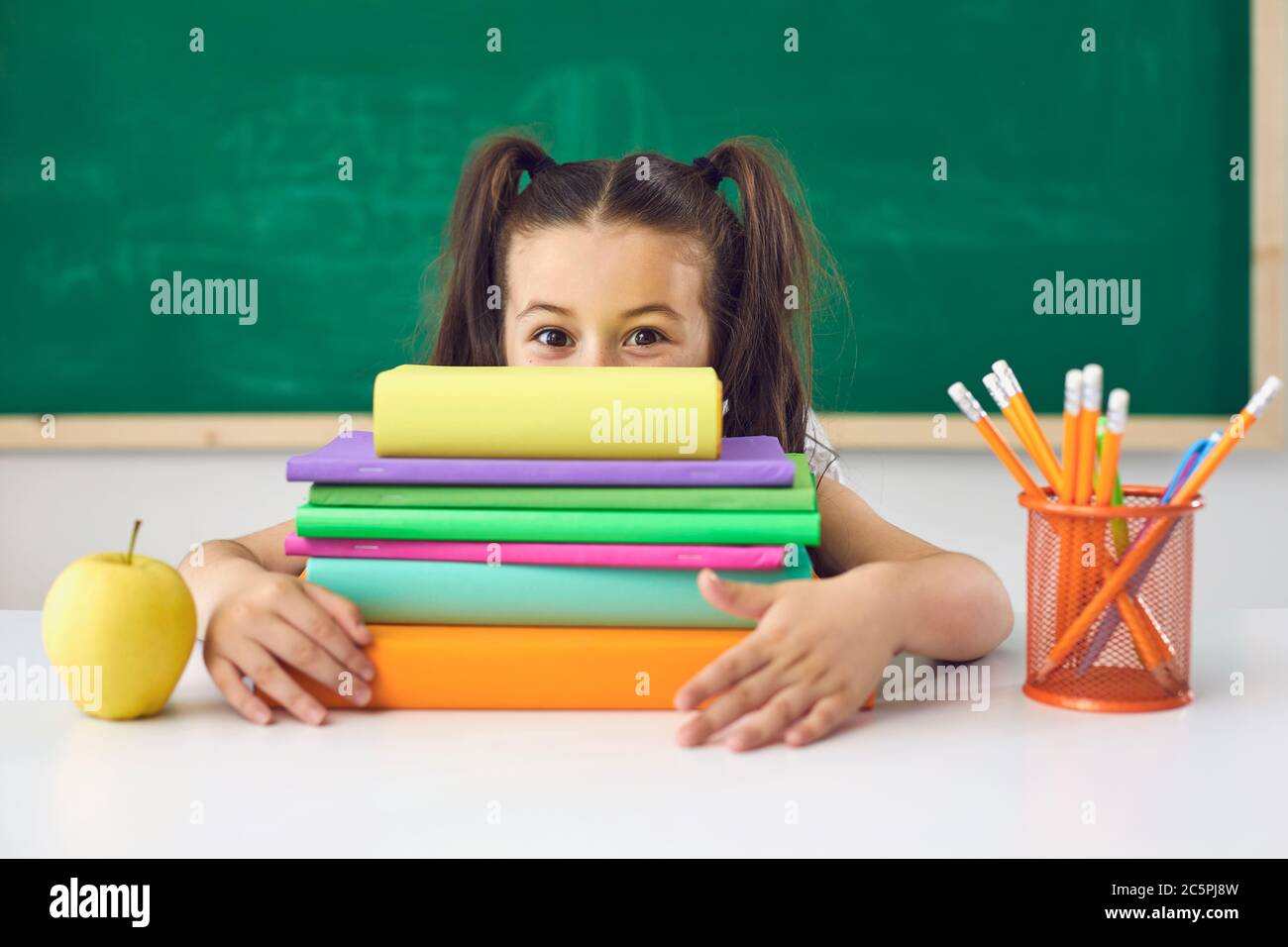 Lustige kleine Schülerin hält ein Buch über ihren Kopf, während in einer Lektion in der Schule sitzen. Stockfoto