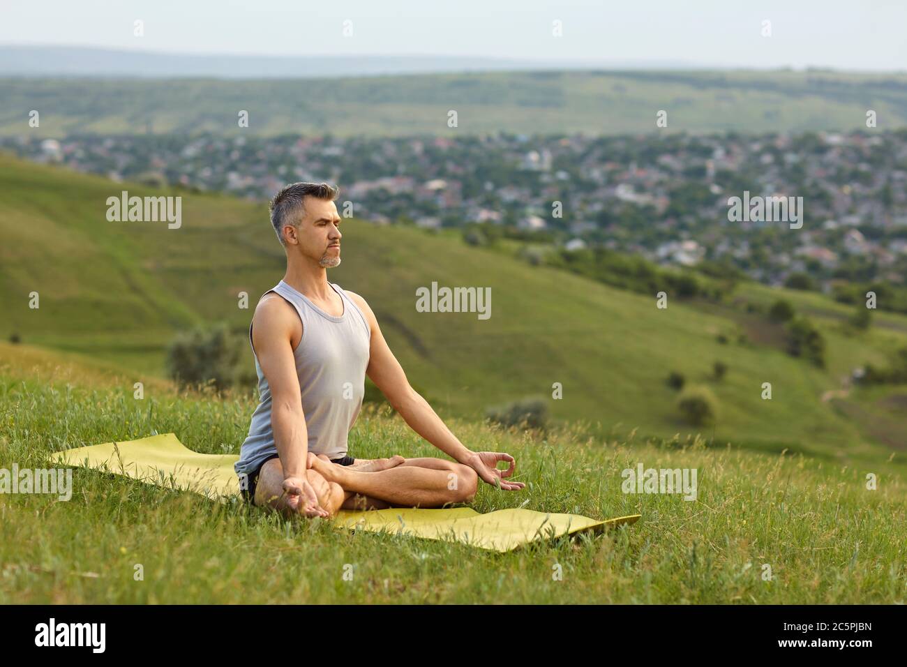 Ein bärtiger, nicht junger Mann praktiziert Yoga-Meditation, sitzt in einer Lotusposition auf dem Gras auf der Sommernatur. Stockfoto