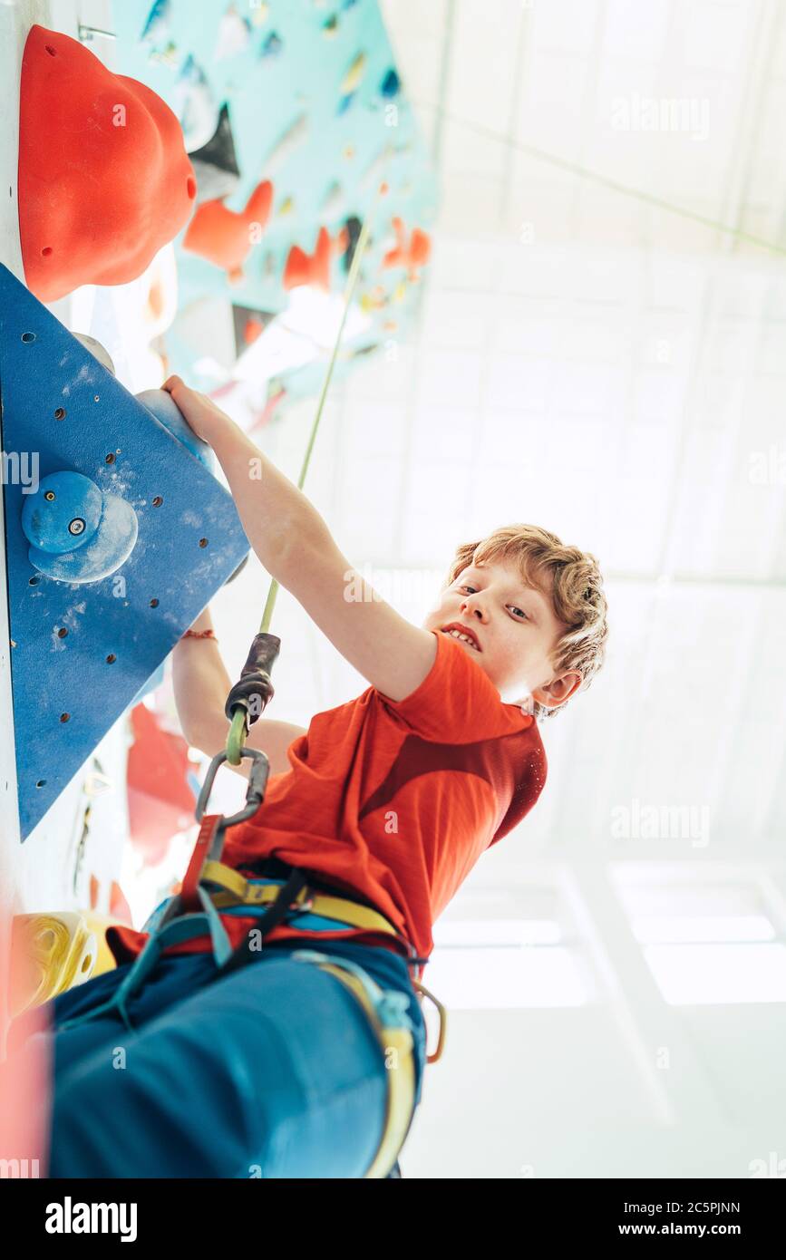Lächelnder Teenager Junge in der Halle der Kletterwand. Boy klettert mit einem Top-Seil, Kreidebeutel und Klettergurt. Aktive Teenager Zeit Ausgaben Konz Stockfoto