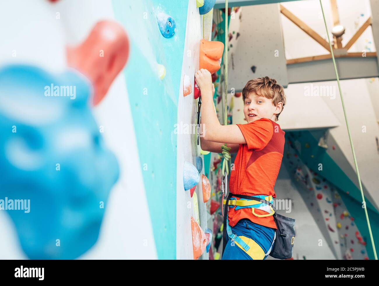 Lächelnder Teenager Junge in der Halle der Kletterwand. Boy klettert mit einem Top-Seil, Kreidebeutel und Klettergurt. Aktive Teenager Zeit Ausgaben Konz Stockfoto
