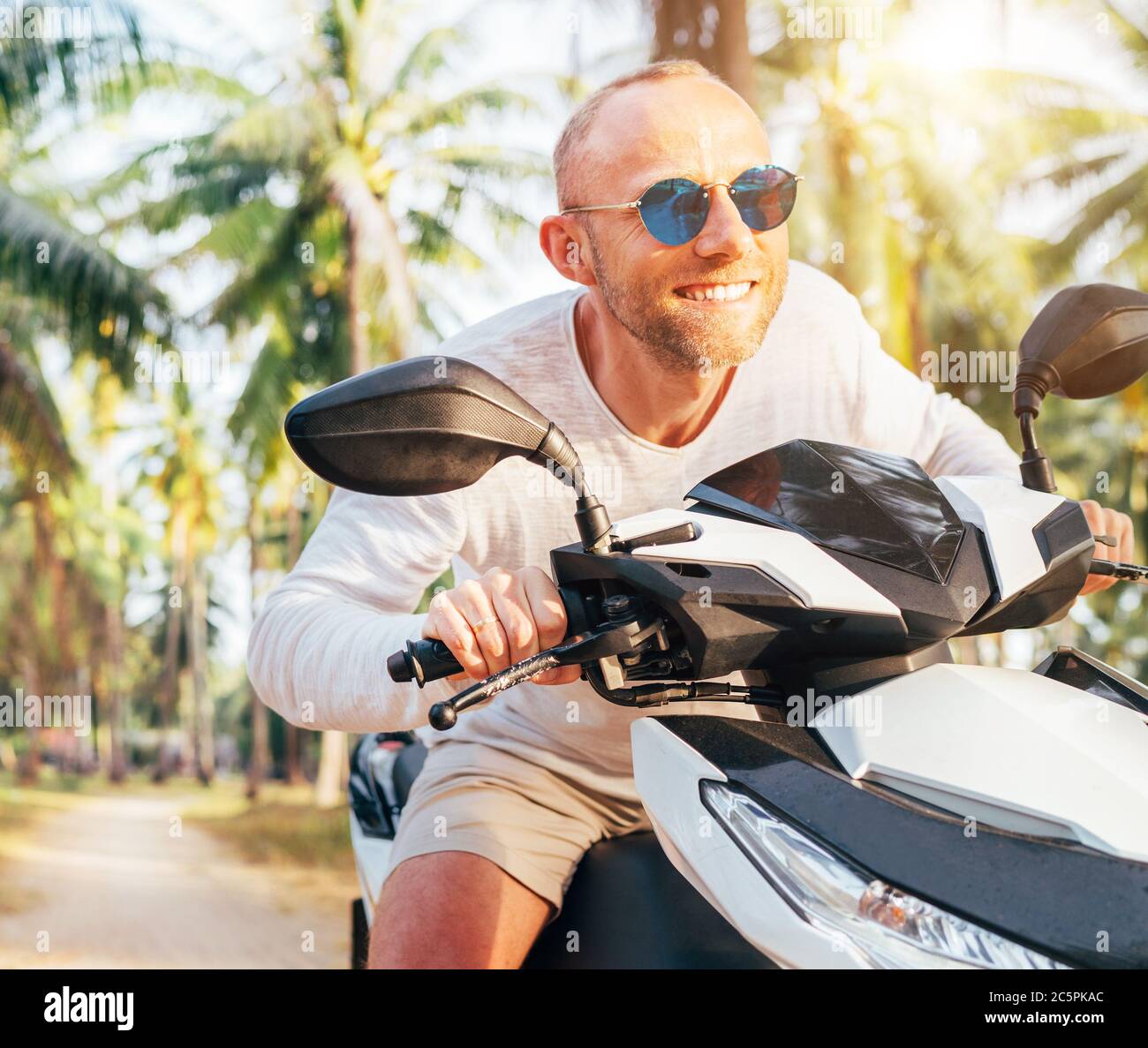 Glücklich lächelnd männlichen Touristen in Sonnenbrille Reiten Motorrad Roller während seines tropischen Urlaubs unter Palmen. Stockfoto
