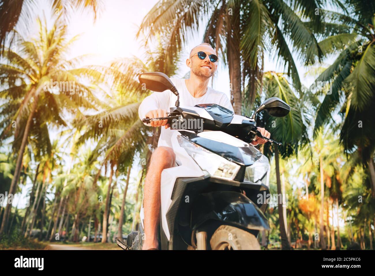 Glücklich lächelnder männlicher Tourist in Sonnenbrille, der während seines tropischen Urlaubs unter Palmen auf Ko Samui Island, Thailand, Motorrad Scooter reitet Stockfoto
