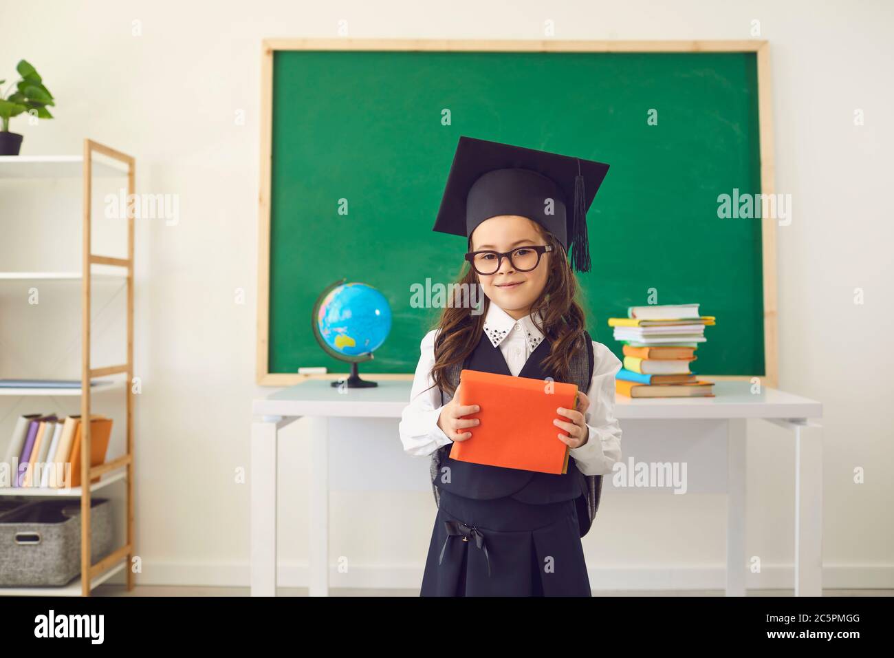 Zurück zur Schule. Ein nettes Schulmädchen in einem Abschlusshut mit Büchern steht vor dem Hintergrund einer Klasse in einer Schule. Stockfoto