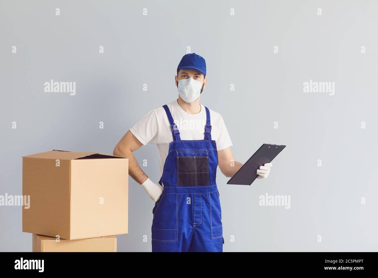 Der Lieferer trägt eine medizinische Maske mit Pappkartons auf grauem Hintergrund. Kurier liefert Online-Bestellung Stockfoto