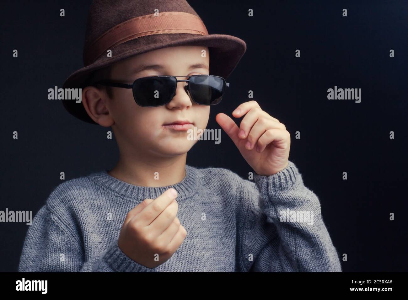 Studio-Portrait eines Jungen in einem grauen Pullover, brauner Hut und Sonnenbrille, auf schwarzem Hintergrund Stockfoto