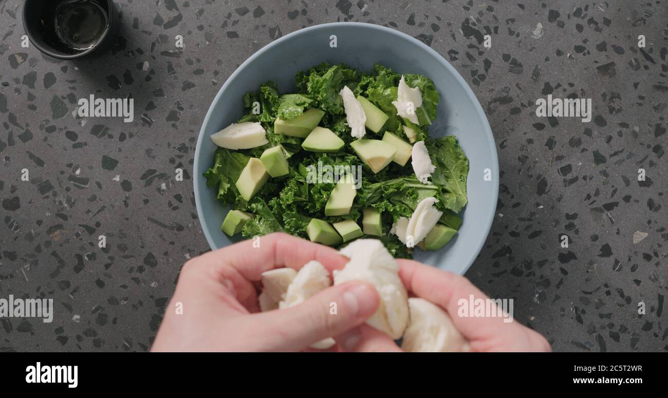 Draufsicht Mann Hände machen Salat setzen Mozzarella über Avocado Und Grünkohl Blätter in blauer Keramik Schüssel auf Beton Hintergrund Stockfoto