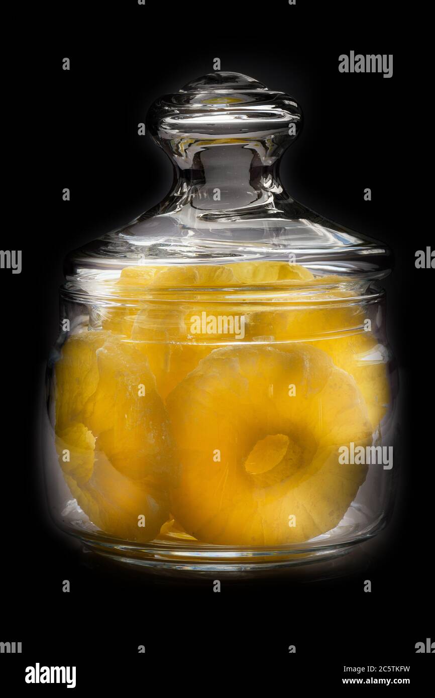 Getrocknete Ananas-Bonbonringe, in Puderzucker, Nahaufnahme unter Sonnenlicht, in einem durchsichtigen Glasglas auf schwarzem Hintergrund liegend. Stockfoto
