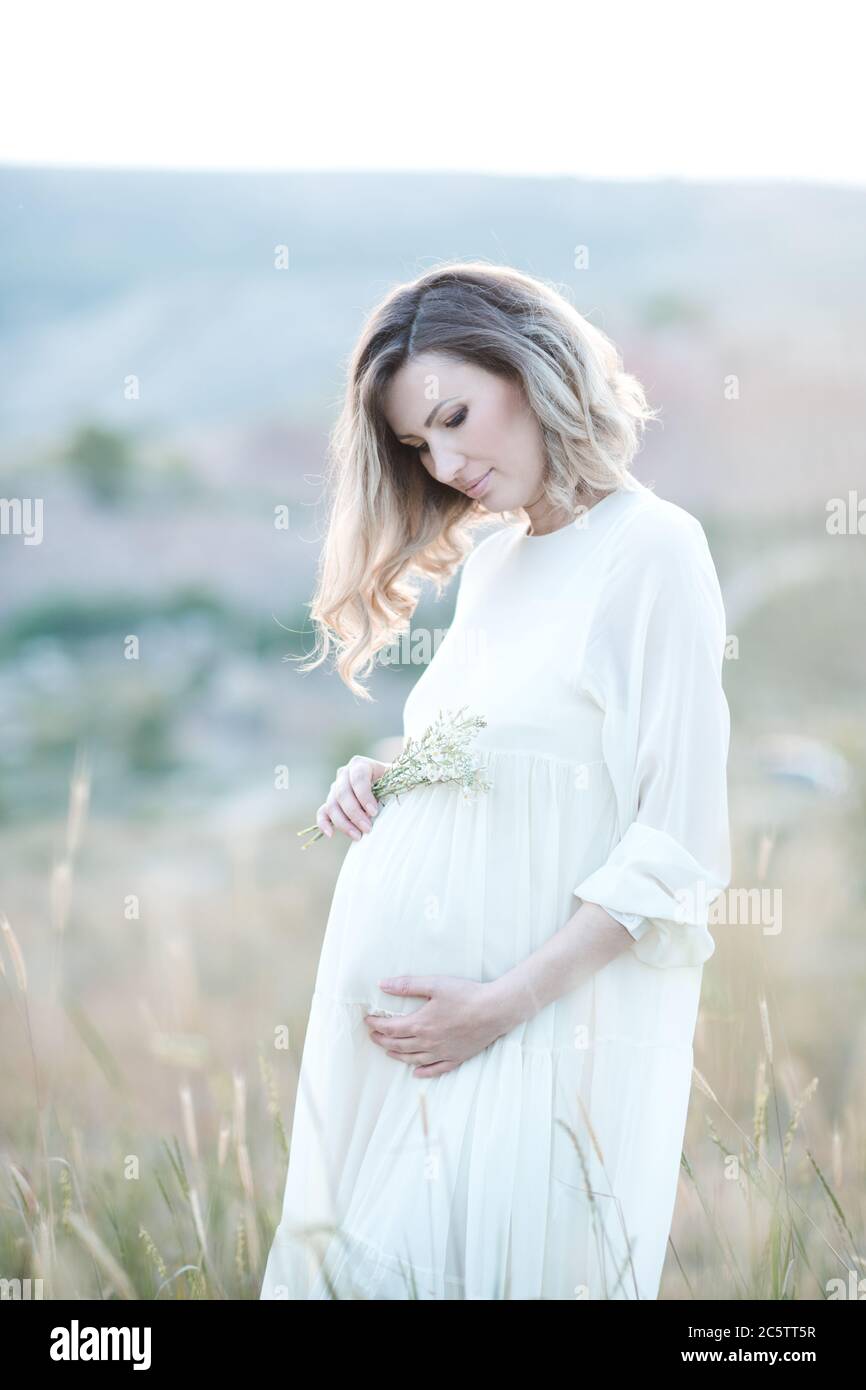 Schöne schwangere Frau trägt weißes Kleid posiert in Wiese im Freien über Sonnenlicht bei Sonnenuntergang. Mutterschaft. Mutterschaft. Stockfoto