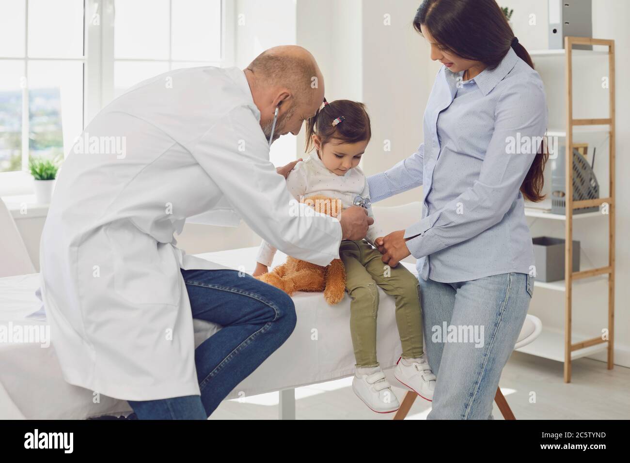 Oberarzt mit Stethoskop hört einem kleinen Mädchen mit Mutterpatienten im Krankenhaus zu. Stockfoto