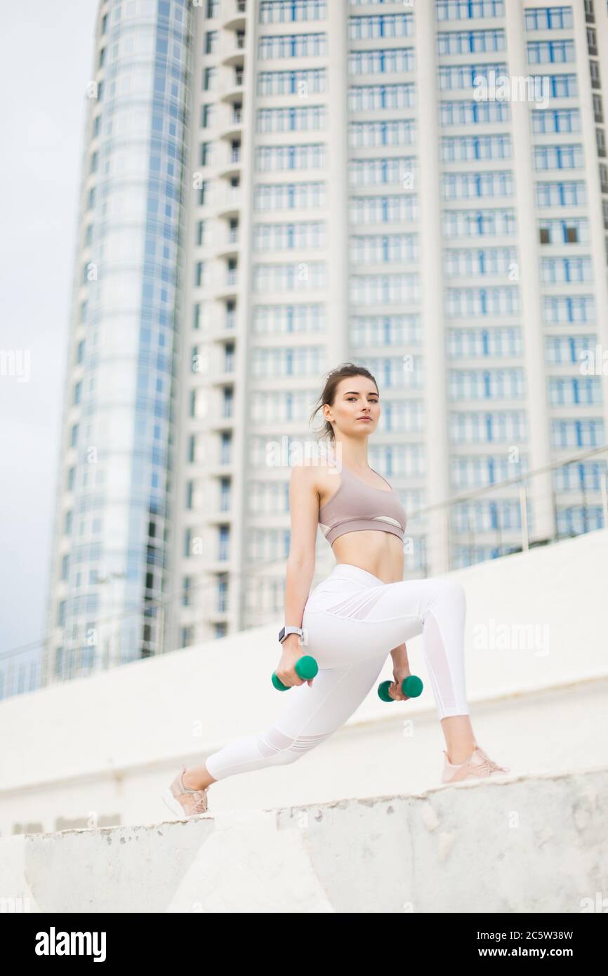 Junge schöne Frau in sportlichen Top und weißen Leggings macht Übungen mit Hanteln in den Händen, während nachdenklich in der Kamera mit Wolkenkratzer auf Stockfoto