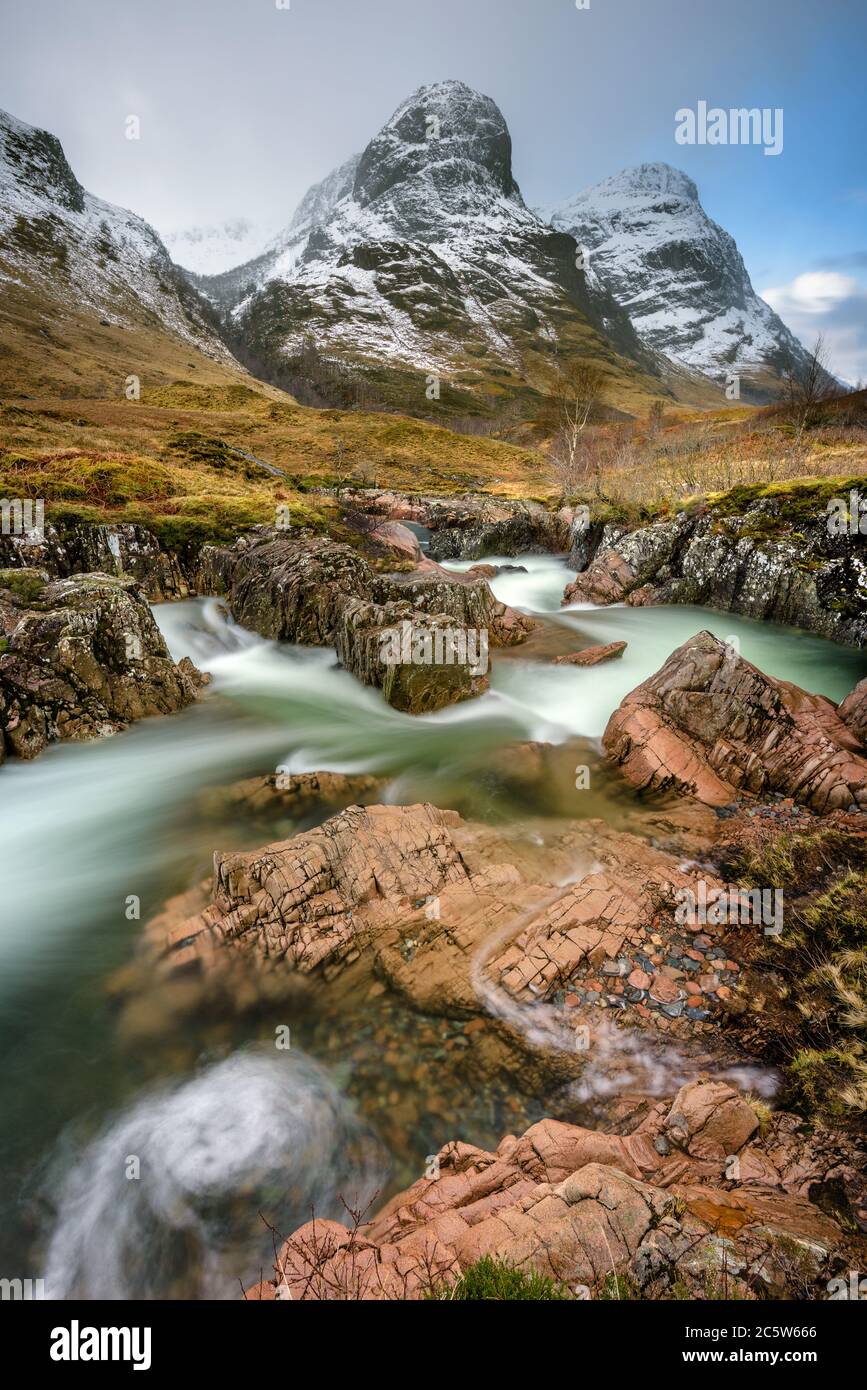 Die Bergkette der Three Sisters in Glencoe ist mit Winterschnee bedeckt und im Vordergrund ein interessanter felsiger Bach. Schottische Highlands, Großbritannien. Stockfoto