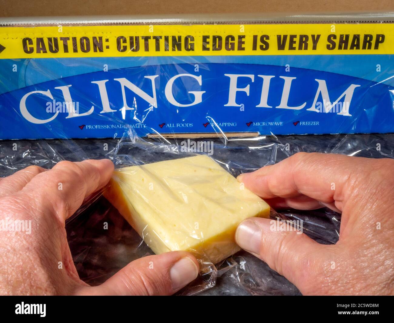 Menschenhände wickeln Frischhaltefolie um ein Stück Käse. Die Abreißrolle dahinter weist darauf hin, dass die gezackte Schneidkante sehr scharf ist. Stockfoto