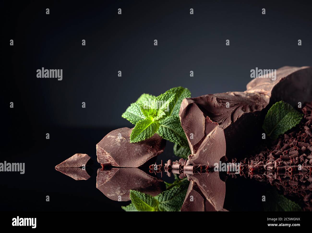 Stücke von Bitterschokolade und Schokoladenstückchen mit Minze auf schwarzem reflektierendem Hintergrund. Speicherplatz kopieren. Stockfoto