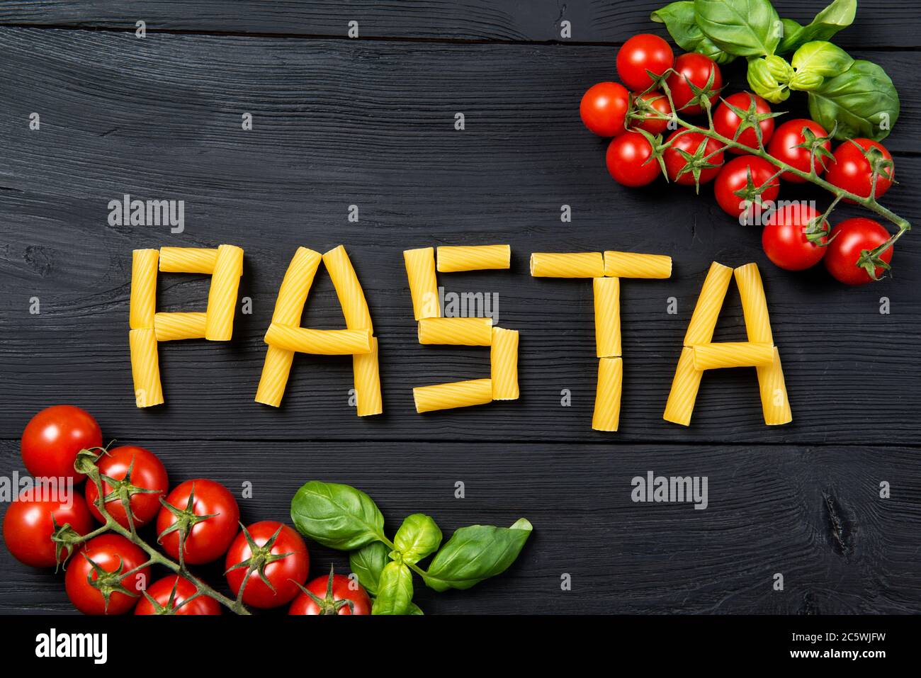 Wortpasta aus Tortiglioni Pasta Macaroni auf dem schwarzen Holztisch, reife Kirschtomaten und Basilikumblätter, Draufsicht Stockfoto