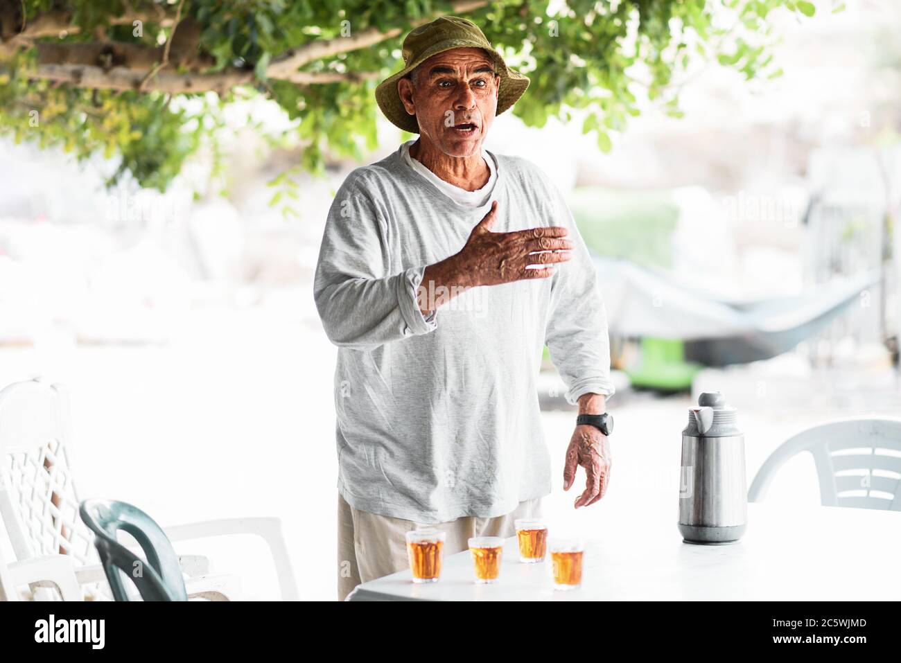 Gesunder aktiver älterer Mann, der im Freien redet und gestikuliert. Arabischer alter Mann streitet mit seinen Freunden, die neben dem Tisch mit traditionellem Tee stehen. Stockfoto