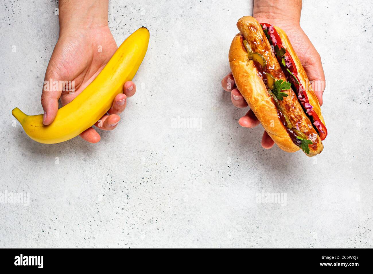 Auswahl gesunder Lebensmittel und ungesunde Lebensmittel Konzept. Vegan vs. Fleischgericht. Banane und Hot Dog in männlichen Händen. Stockfoto