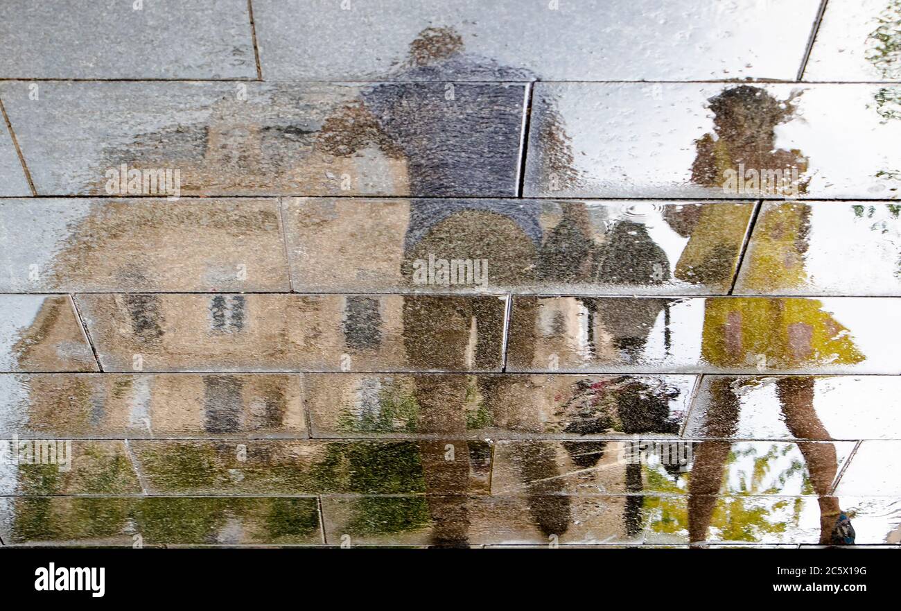 Verschwommene Spiegelschatten Silhouette eines Mannes und einer Frau in einem gelben Kleid zu Fuß auf einer nassen Straße Pfütze in der Fußgängerzone der Stadt auf einem regnerischen und Stockfoto