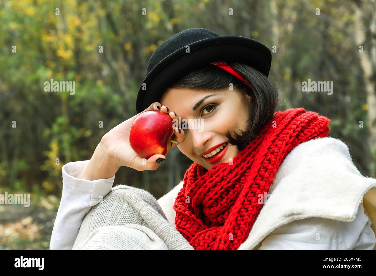 Positive Frau in schwarzem Hut und rotem Schal mit einem roten Apfel in der Hand lächelt und schaut auf die Kamera. Stockfoto