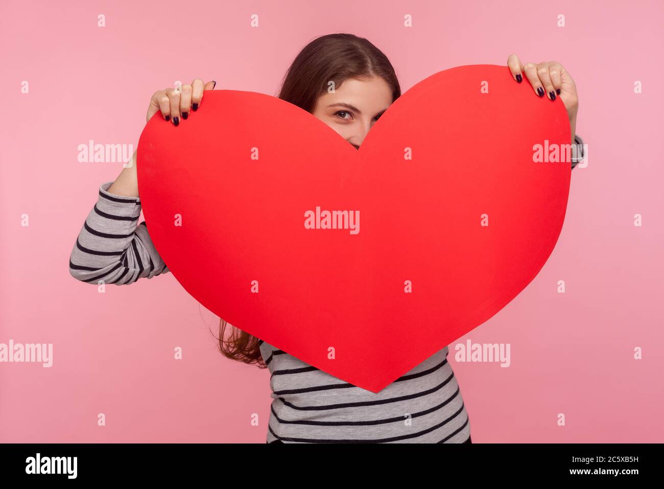 Sei mein Valentinstag! Portrait von kokettiert Frau piepend aus riesigen roten Papier Herz mit verspielten glücklich Blick, zeigt Liebe Pflege, romantische Gefühle. ich Stockfoto