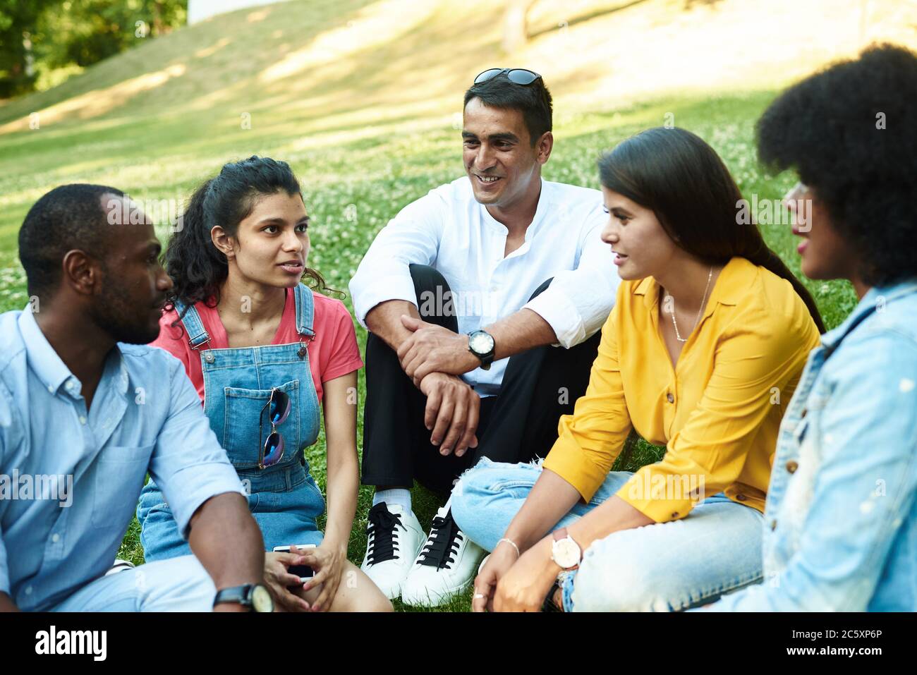 Afghanische, schwarze, mexikanische und indische Plauderei, während sie im Park auf dem Gras sitzen. Das Konzept der Kommunikation verschiedener Nationalitäten. Verschiedene Menschen und Freunde. Stockfoto