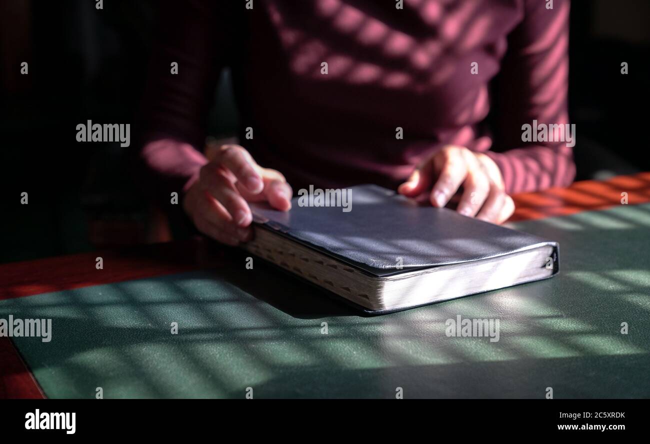 Frauenhand auf der Heiligen bibel, Fokus auf das Buch. Schatten der Fensterläden erzeugt Linien und Muster. Stockfoto