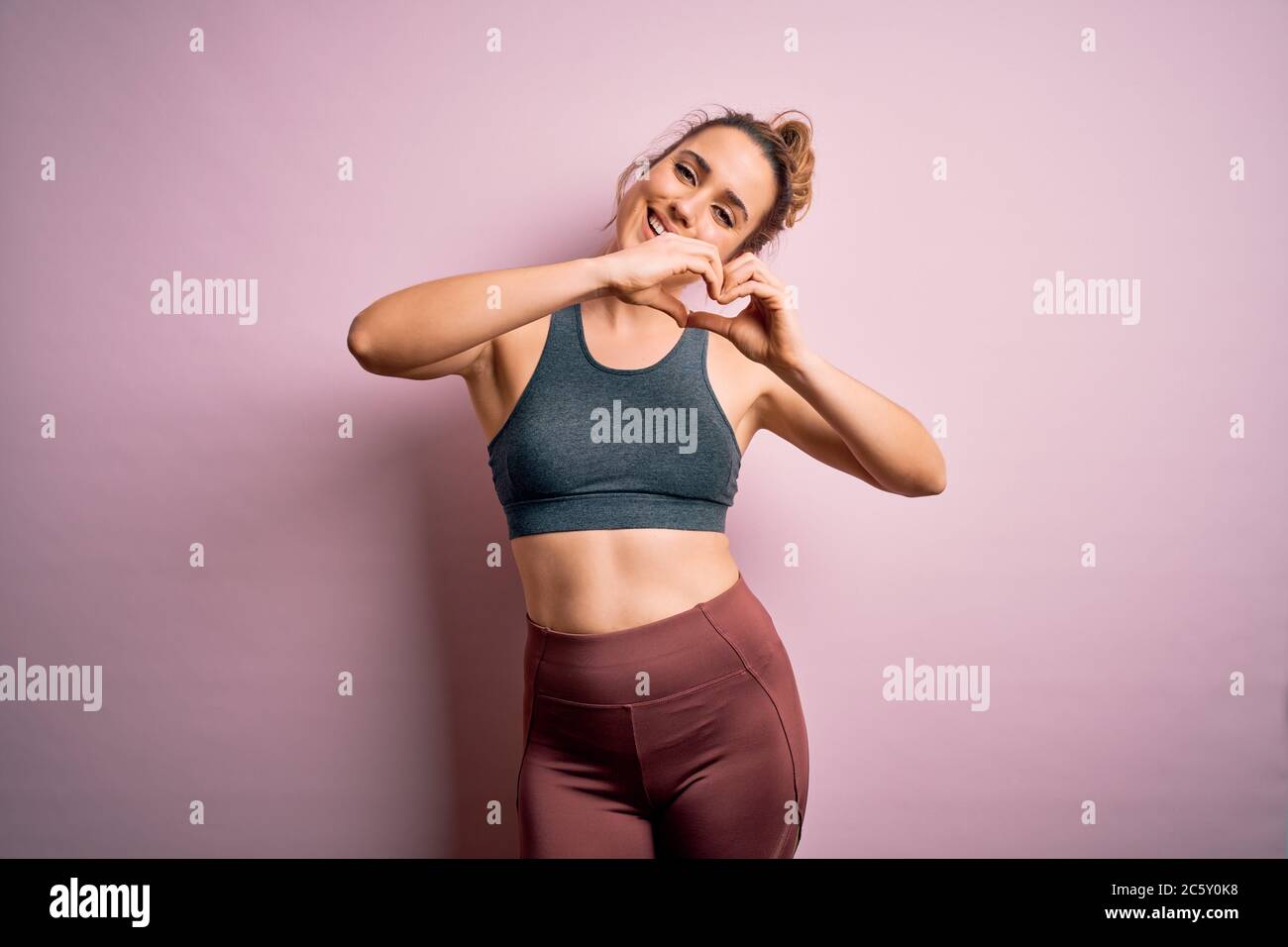 Junge schöne blonde Sportlerin Sport Sport Sport tragen Sportbekleidung über rosa Hintergrund lächelnd in Liebe tun Herz Symbol Form mit Händen. Romantisches c Stockfoto