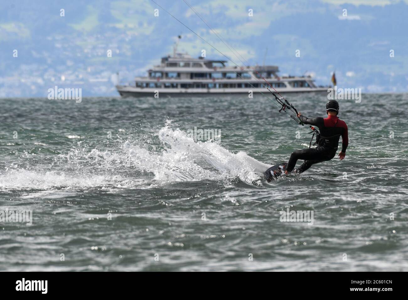 Friedrichshafen, Deutschland. Juli 2020. Ein Kitesurfer jagt seinen Drachen in Böen bis zu 40 Stundenkilometer über den Bodensee, während im Hintergrund ein Passagierschiff der Weißen Flotte vorbeifährt. Dahinter ist die Schweiz zu sehen. Quelle: Felix Kästle/dpa/Alamy Live News Stockfoto