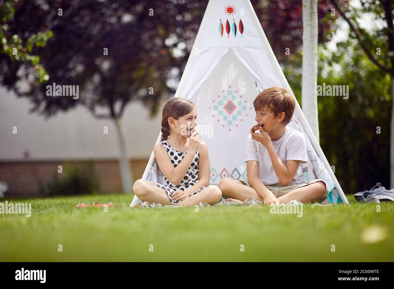 Nette glückliche kleine Kinder sitzen im Tipi im Hinterhof und isst Kekse. Stockfoto