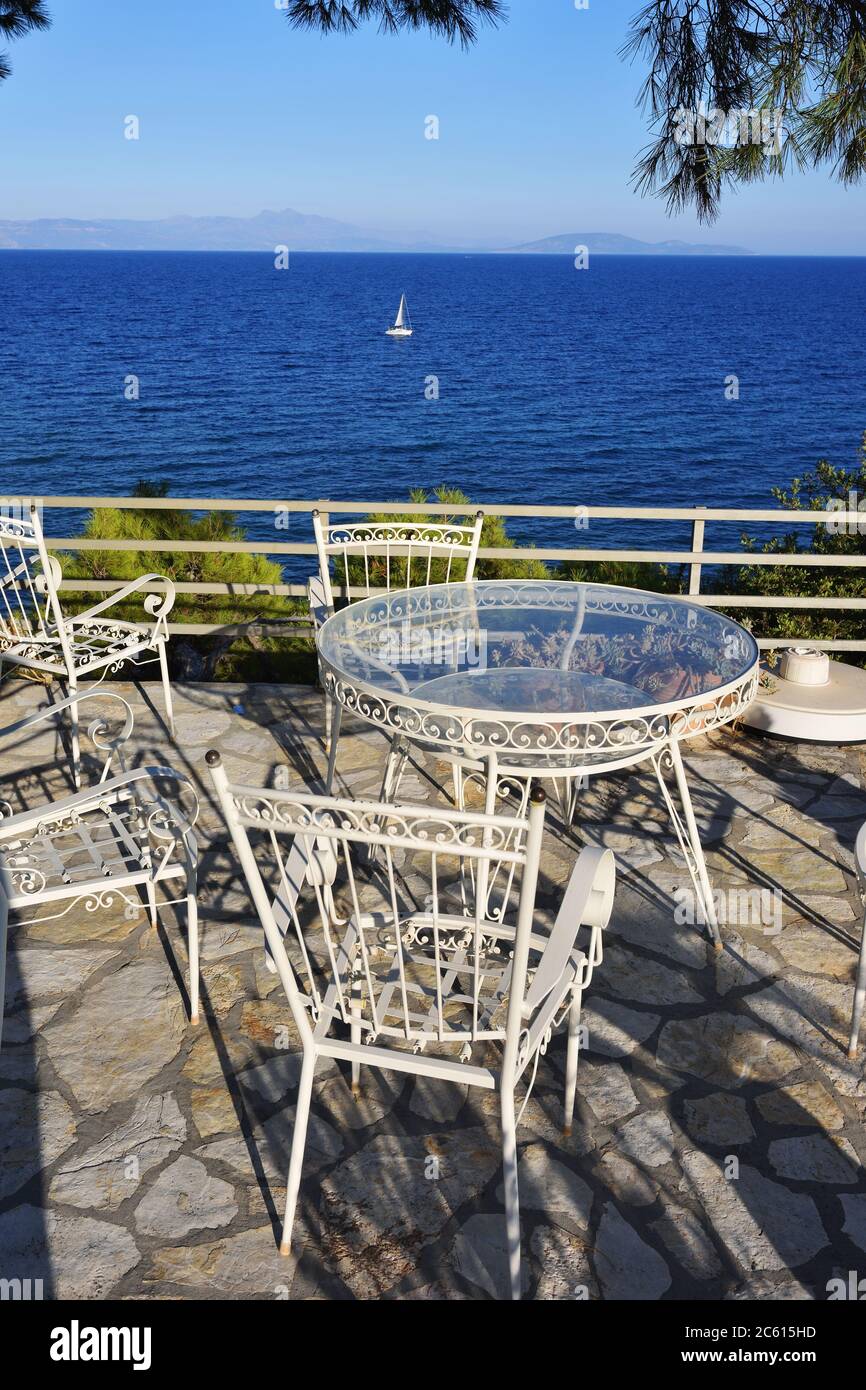 Picknicktisch und Stühle unter Pinien im Innenhof an der Küste der Ägäis bei Sonnenuntergang, weiße Segelyacht im Hintergrund. Warmes Abendlicht. A Stockfoto