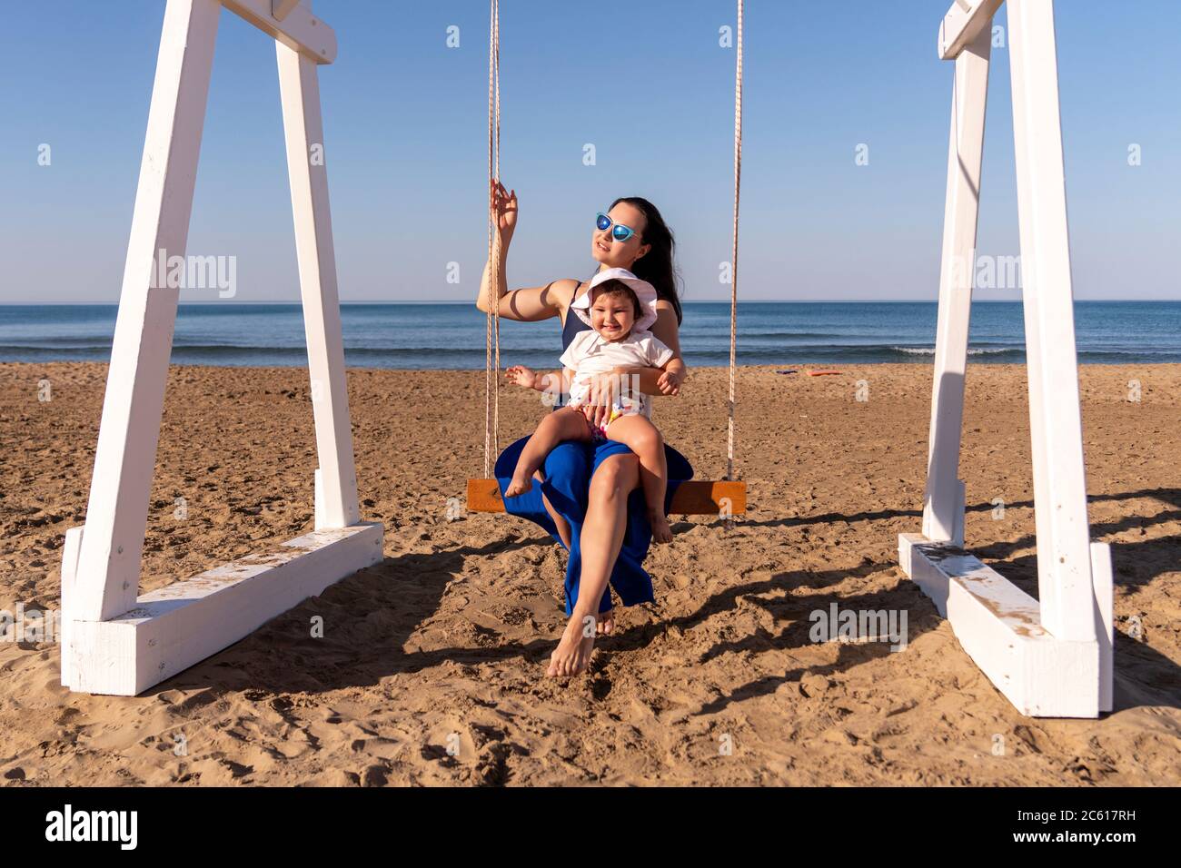 Schöne Frau schwingt mit ihrem Baby am feinen Sandstrand und genießt die idyllische Szene von Sonne und Meer. Wunderschöner Sandstrand in Antalya Stockfoto