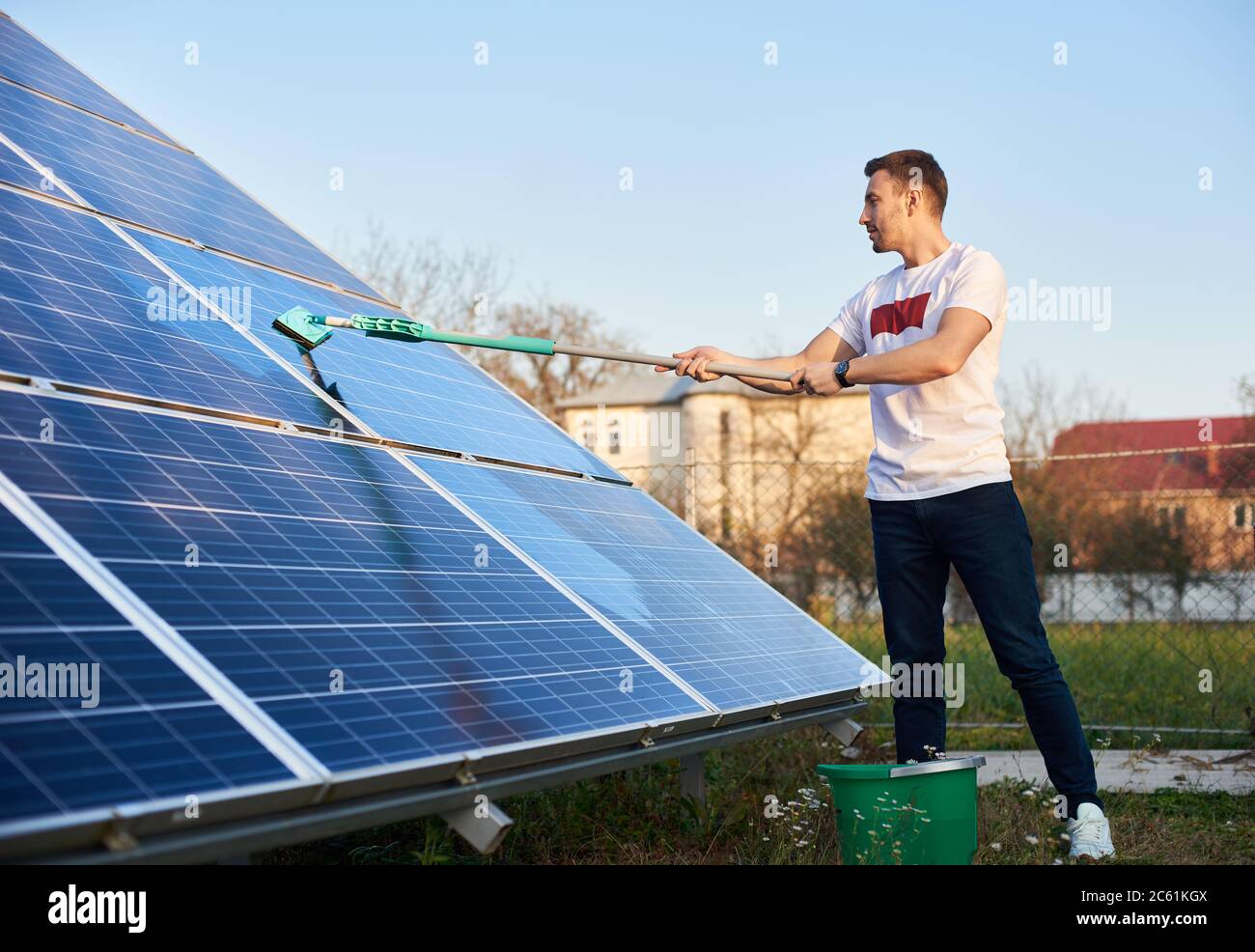 Junger Mann putzt Solarpanel auf einem Grundstück in der Nähe des Hauses. Der Typ ist in der Nähe stilvoll gekleidet und hat einen Eimer Wasser Stockfoto