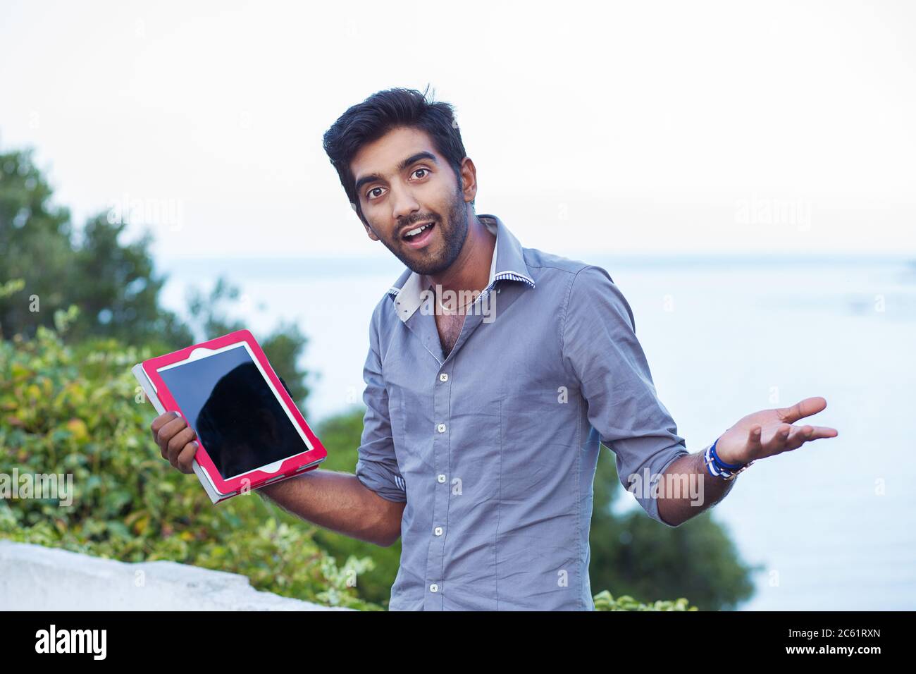Ein lächelnder Mann, Student zeigt Gleichgültigkeit, also was Geste mit Händen halten zeigt einen Tablet-Pad-Bildschirm, während vor einem Stadtpark Witz stehen Stockfoto