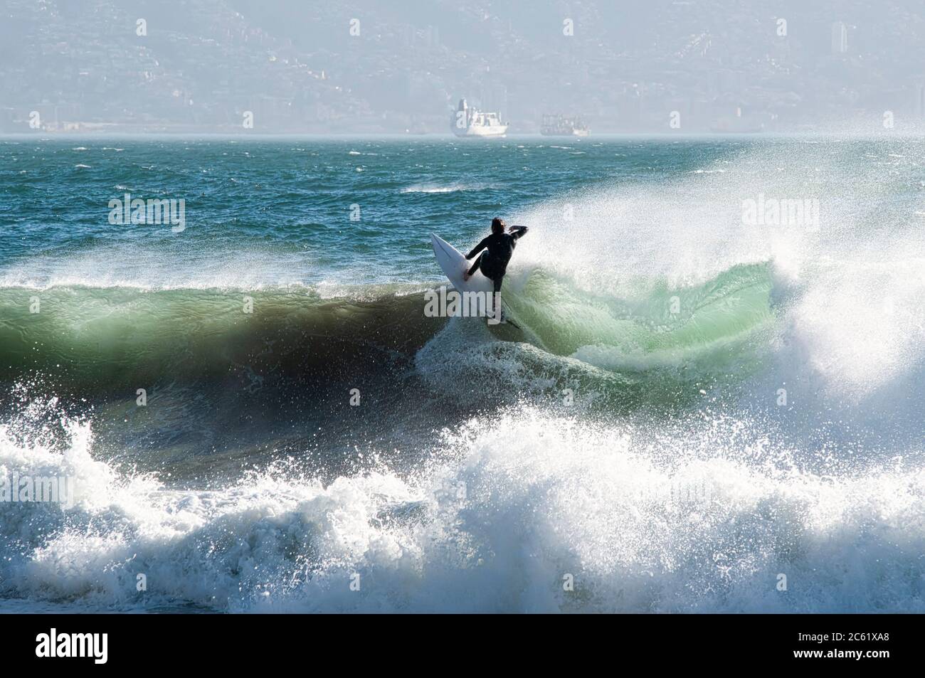 Surfer auf dem Kamm der Welle, vom Strand Reñaca aus gesehen, Zentralküste Chiles. Im Hintergrund ein Schiff, das im Hafen von Valparaiso ankommt Stockfoto