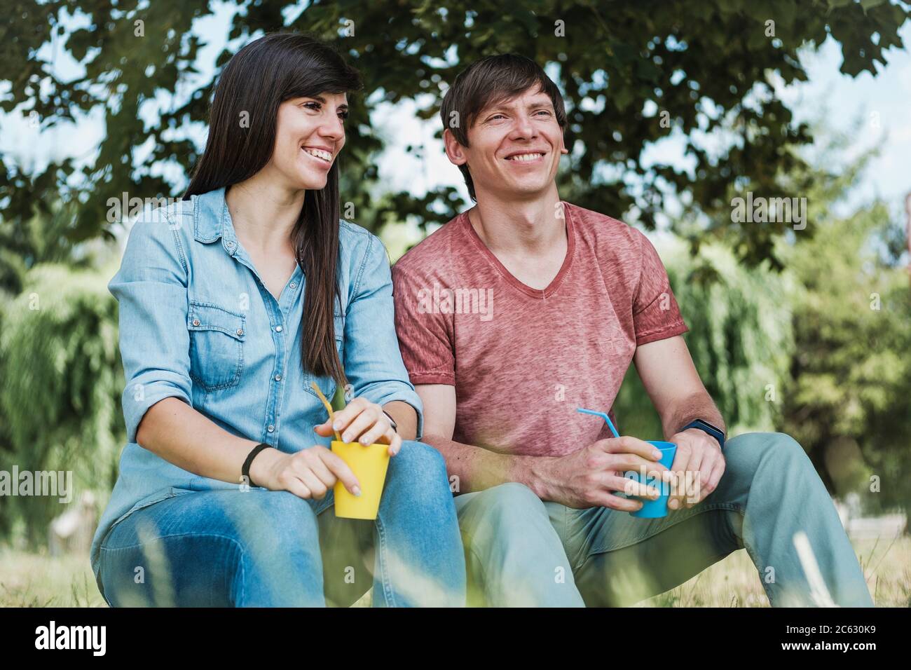 Junge glückliche Paar entspannen im Schatten eines Baumes mit bunten Tassen Saft in einem gesunden aktiven Lebensstil Konzept Stockfoto