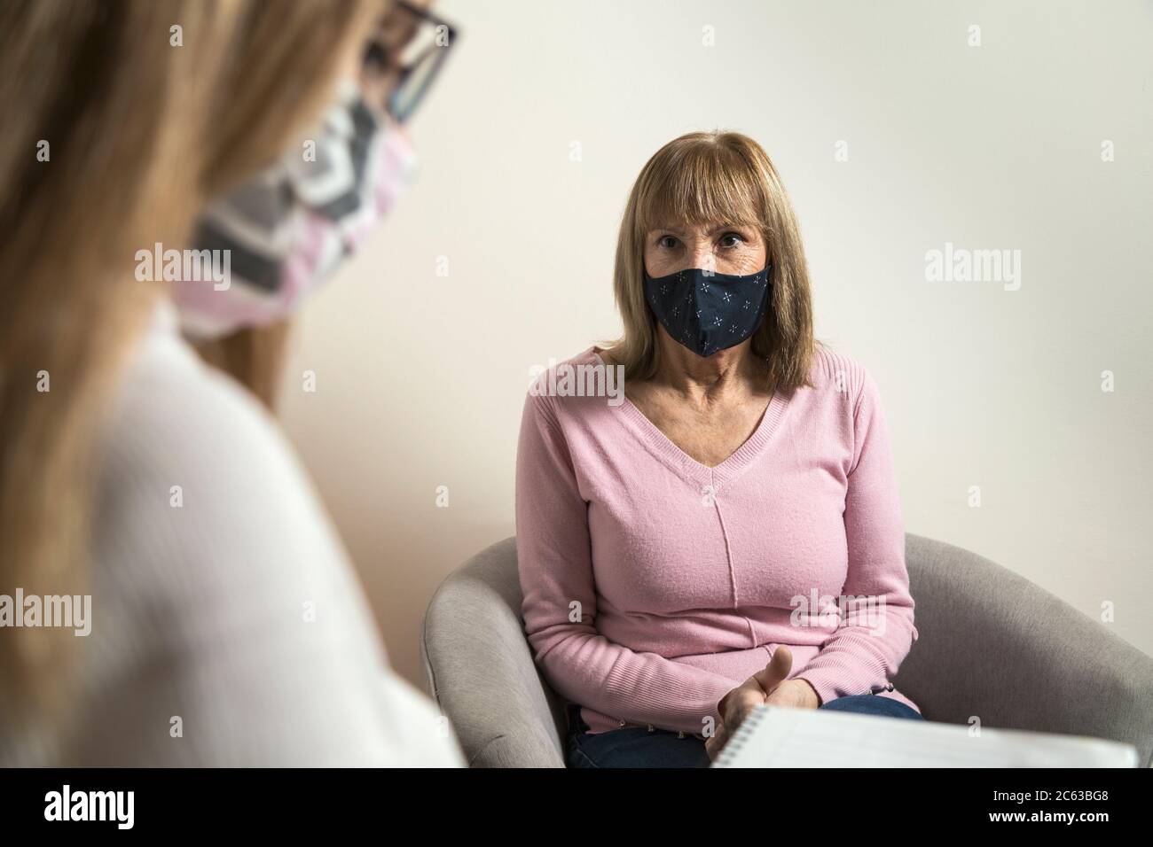 Frau mit Kinnriemen im Gespräch mit einer anderen, die Notizen in einem Notizbuch. Beide mit medizinischen Masken. Aufmerksamkeit Protokoll.Sorgen Frau, die von betreut Stockfoto